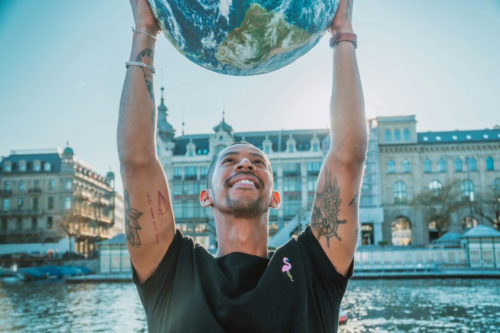 笑顔で地球の模型を掲げる男性。背景にはヨーロッパ風の建物と水辺の風景。